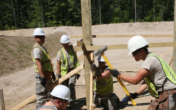 A group of five military personnel in camouflage uniforms are engaged in construction work at an outdoor site. They are erecting wooden framing structures on a sandy, cleared ground area surrounded by a treeline. One worker in an orange safety vest is kneeling to stabilize a post while another swings a sledgehammer to drive it into the ground.