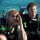 Two men wearing full scuba diving equipment are positioned near a body of water, appearing to be in a post-dive or pre-dive situation. Both are equipped with tanks, BCDs (buoyancy control devices), regulators, and hoses. The man in the foreground has blonde hair and is raising his hand toward his face, while the man behind him wears a black wetsuit and tactical gloves.