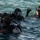 Three divers in full scuba gear are positioned in open water, appearing to be conducting a briefing or equipment check at the surface. The diver on the right, wearing a simple dive mask and mustache, appears to be communicating or signaling with a raised hand holding an object. The equipment and tactical appearance suggest this may be a military or professional dive operation.