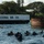 A group of scuba divers are in the water near the wreck site of the USS Arizona (BB-39) at Pearl Harbor, Hawaii. The white marker buoy or platform clearly displays the ship's designation 'USS ARIZONA BB 39', with a portion of the rusted wreck visible above the waterline to the right. The divers appear to be conducting an underwater inspection or documentation dive at this historic memorial site.