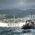 A rigid inflatable boat (RIB) carrying multiple military personnel in tan/khaki uniforms is underway on the water at Pearl Harbor, Hawaii. In the background, the distinctive white USS Arizona Memorial structure is clearly visible, along with an American flag on a flagpole. The mountainous terrain of Oahu and a residential hillside community are visible in the far background under overcast skies.