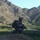 A U.S. soldier in full combat gear is crouched on an elevated dirt mound, observing a valley below containing terraced agricultural fields and a small village. The backdrop features rugged, arid mountain terrain with sparse vegetation, consistent with Central Asian or South Asian highland geography. The scene appears to depict a reconnaissance or security overwatch position during a military patrol.