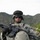 A U.S. Army soldier in full combat gear is photographed in a mountainous terrain, likely during a deployment. The soldier is wearing ACU (Army Combat Uniform) pattern camouflage, a helmet with a black cover, and is holding an M4 carbine rifle. The rugged, arid mountain landscape visible in the background is consistent with terrain found in Afghanistan or similar regions.