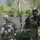 Two U.S. Army soldiers in full combat gear are positioned near a fast-moving river in a mountainous, vegetated terrain. Both wear ACU (Army Combat Uniform) pattern camouflage, helmets, and tactical vests with backpacks. The soldier on the right is facing the camera holding a rifle, while the soldier on the left faces away toward the river.