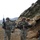 The image shows a group of soldiers in U.S. Army ACU (Army Combat Uniform) camouflage conducting a patrol along a rocky mountain trail. The terrain and environment are consistent with a mountainous region, likely Afghanistan based on the landscape and operational context. The soldiers are fully equipped with weapons, body armor, and tactical gear.