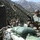 Military personnel are positioned at a fortified outpost in a mountainous terrain, manning what appears to be an autocannon or heavy weapon system. The position is reinforced with green sandbags and features a camouflage-covered structure. The rugged, heavily vegetated mountain range in the background suggests a high-altitude combat or forward operating base environment.