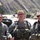 Three U.S. Army soldiers in full combat gear pose together at what appears to be a forward operating base in a mountainous region, likely Afghanistan. The soldiers are wearing Army Combat Uniforms (ACU) with tactical vests and equipment. A rugged mountain range and military vehicles are visible in the background.