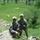 Two U.S. Army soldiers in ACU (Army Combat Uniform) pattern camouflage pose on a rocky hillside overlooking a lush green valley. Both wear Advanced Combat Helmets and carry M4-style carbines. A third soldier is visible in the background standing in the field, and an orange panel marker is visible on the ground in the valley.
