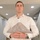 A young adult male in a white dress shirt stands indoors holding a folded American flag in the traditional triangular military presentation fold. His expression is solemn and serious, suggesting a ceremonial or commemorative context. The setting appears to be a residential interior with recessed ceiling lighting.
