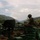 A soldier in camouflage uniform and helmet is positioned at a fortified fighting position constructed with green sandbags, overlooking a mountainous valley. The image is date-stamped July 12, 2008, suggesting a combat or observation post in a mountainous region. Various military equipment including what appears to be a crew-served weapon and mortar rounds are visible at the position.