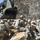 Military personnel in U.S. Army combat uniforms are conducting a MEDEVAC or casualty evacuation operation in a rocky, mountainous terrain. A CH-47 Chinook helicopter is visible overhead or nearby, suggesting an active or imminent medical evacuation. Soldiers are gathered around what appears to be a litter/stretcher with a casualty, positioned behind a sandbag fortification.