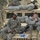 Three U.S. Army soldiers in ACU (Army Combat Uniform) are resting on and around a tan HUMVEE military vehicle marked with 'ISAF' insignia, indicating deployment with the International Security Assistance Force, likely in Afghanistan. Two soldiers are seated on the hood of the vehicle while a third is lying on the roof. The scene depicts a rest period during a military operation in a rocky, arid environment.