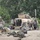 The image shows a group of military personnel in desert camouflage uniforms gathered around a tan/beige HMMWV (Humvee) in what appears to be a forward operating area. Soldiers are engaged in various tasks including vehicle maintenance, equipment staging, and rest. The setting appears to be a rural or semi-arid environment with dense tree cover in the background.