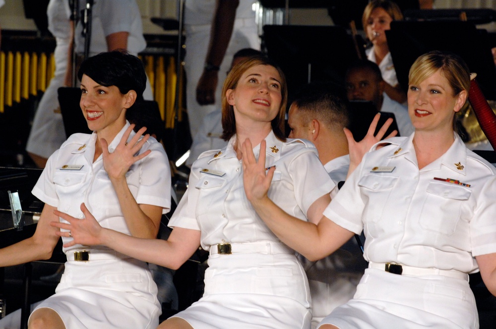 Three women in white military dress uniforms are seated together, smiling and gesturing with their hands, appearing to be performing or participating in a musical or ceremonial event. They wear matching white short-sleeve uniforms with gold belt buckles, name tags, and insignia. A band or orchestra is visible in the background.