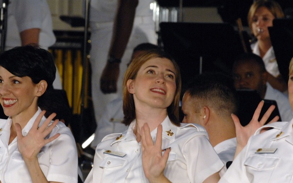 Three women in white military dress uniforms are seated together, smiling and gesturing with their hands, appearing to be performing or participating in a musical or ceremonial event. They wear matching white short-sleeve uniforms with gold belt buckles, name tags, and insignia. A band or orchestra is visible in the background.