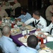 A group of military personnel in various uniforms are seated together at tables in what appears to be a dining facility (mess hall or chow hall). The group includes individuals in Navy white dress uniforms, Marine Corps camouflage utilities, and tan/khaki service uniforms. Meals appear to be mostly finished, suggesting a social dining event or meeting.