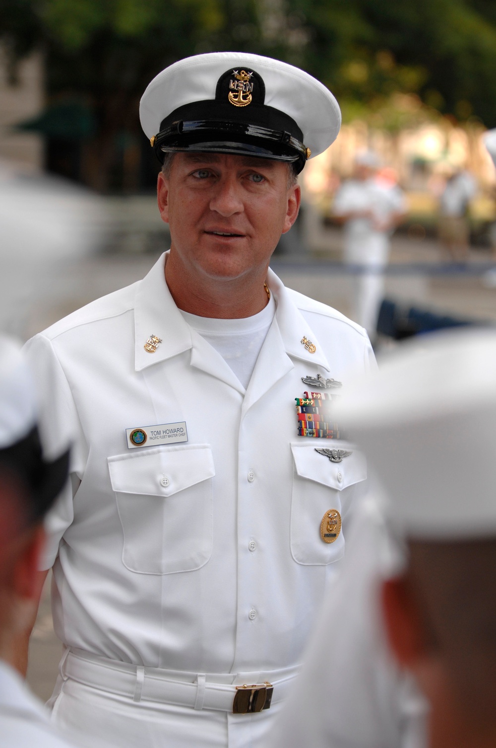 A U.S. Navy senior enlisted sailor in Service Dress White uniform is photographed outdoors at what appears to be a formal military event. His name tag reads 'TOM HOWARD' with the subtitle 'PACIFIC FLEET MASTER CHIEF,' indicating a very senior enlisted position. He wears multiple decorations, warfare badges, and the distinctive Master Chief Petty Officer cap device.