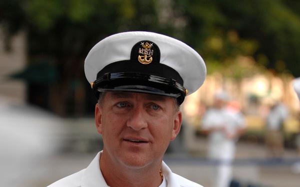 A U.S. Navy senior enlisted sailor in Service Dress White uniform is photographed outdoors at what appears to be a formal military event. His name tag reads 'TOM HOWARD' with the subtitle 'PACIFIC FLEET MASTER CHIEF,' indicating a very senior enlisted position. He wears multiple decorations, warfare badges, and the distinctive Master Chief Petty Officer cap device.