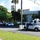 The image shows a U.S. Customs and Border Protection (CBP) vehicle and what appears to be a specialized operations truck parked on a road, with a group of observers gathered on the left side. The setting appears to be a government or institutional facility in a tropical location, likely Hawaii, based on the palm trees and building architecture. This appears to be a demonstration or official event given the assembled crowd of onlookers.