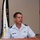 A U.S. Air Force officer wearing a light blue short-sleeve uniform with two-star major general rank insignia is speaking at a wooden podium. His name tag reads 'COMPTON' and he wears pilot wings on his chest. Military flags are visible in the background.
