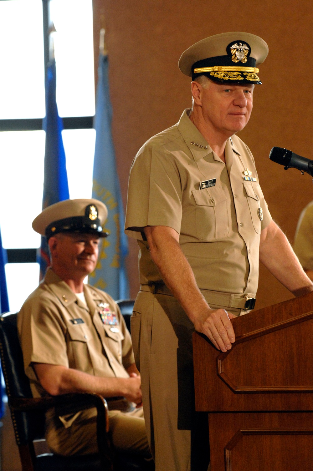 Two U.S. Navy officers in tan service uniform (khaki) are present at what appears to be an official military ceremony or briefing. The senior officer standing at a wooden podium with a microphone has gold 'scrambled eggs' on his cap visor, indicating flag officer (admiral) rank, with rank insignia visible on his collar. A second officer is seated behind him, wearing a cap with a single gold band indicating a lower rank.