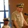 Two U.S. Navy officers in tan service uniform (khaki) are present at what appears to be an official military ceremony or briefing. The senior officer standing at a wooden podium with a microphone has gold 'scrambled eggs' on his cap visor, indicating flag officer (admiral) rank, with rank insignia visible on his collar. A second officer is seated behind him, wearing a cap with a single gold band indicating a lower rank.