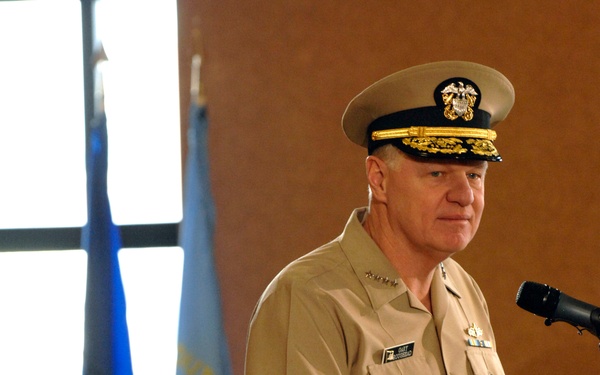 Two U.S. Navy officers in tan service uniform (khaki) are present at what appears to be an official military ceremony or briefing. The senior officer standing at a wooden podium with a microphone has gold 'scrambled eggs' on his cap visor, indicating flag officer (admiral) rank, with rank insignia visible on his collar. A second officer is seated behind him, wearing a cap with a single gold band indicating a lower rank.