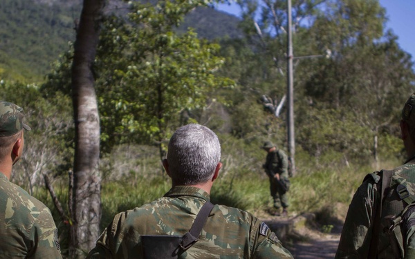 Three soldiers in camouflage uniforms are observed from behind, standing on a dirt path in a wooded, hilly environment. They appear to be observing or monitoring a fourth soldier visible further down the path. The scene suggests a military patrol or field exercise in a forested terrain.