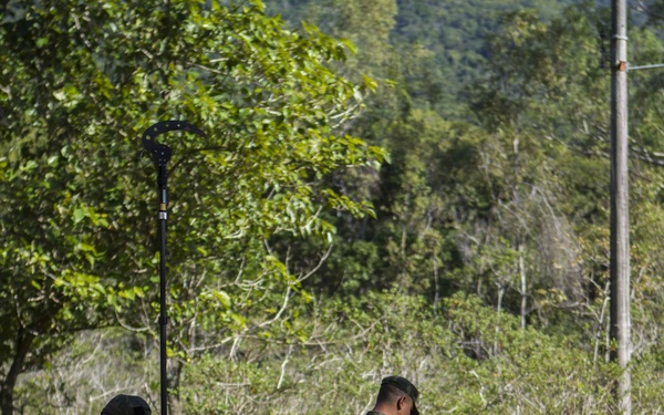 Two individuals in military camouflage uniforms are conducting what appears to be a demining or metal detection sweep along a rural dirt path. One person holds a long pole-mounted device vertically while the other actively sweeps a handheld metal detector close to the ground. The setting is a forested, rural area with a concrete drainage channel visible alongside the path.