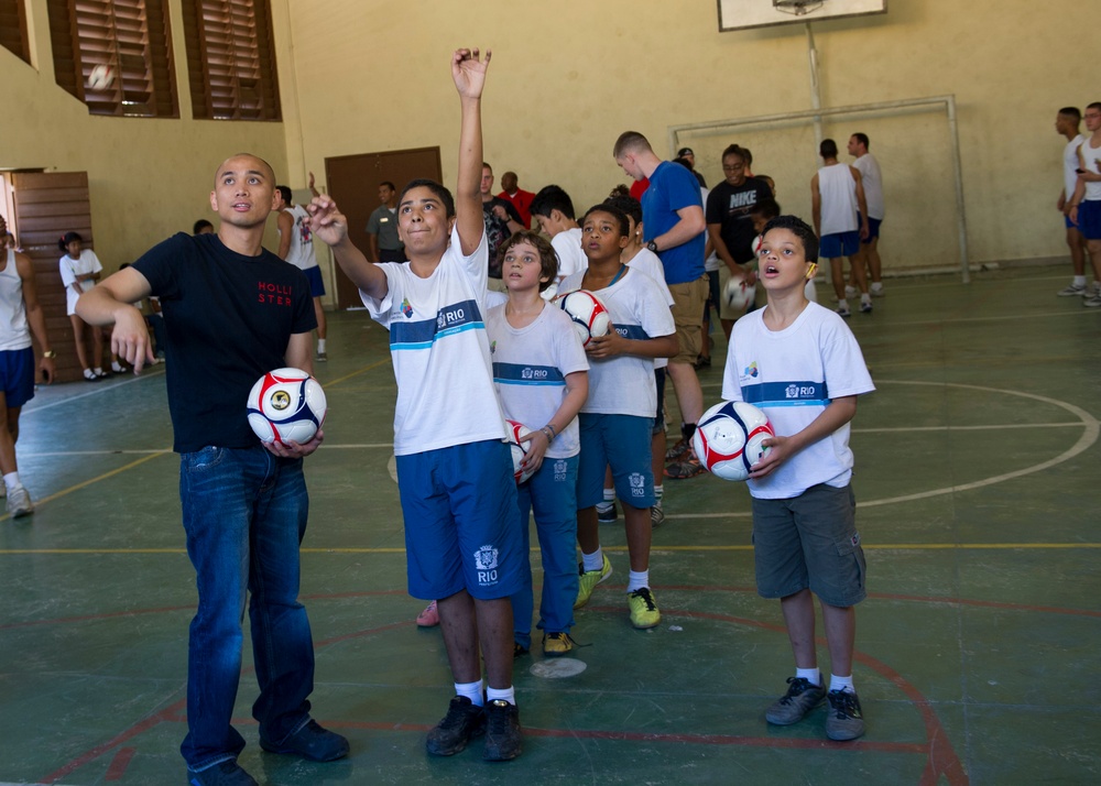 A group of children wearing white 'RIO' branded uniforms are participating in an indoor soccer training session in a gymnasium. An adult instructor in a black Hollister shirt holds a soccer ball while a young boy points upward, apparently tracking a ball or demonstrating a technique. The setting appears to be a community sports program in Rio de Janeiro, Brazil.