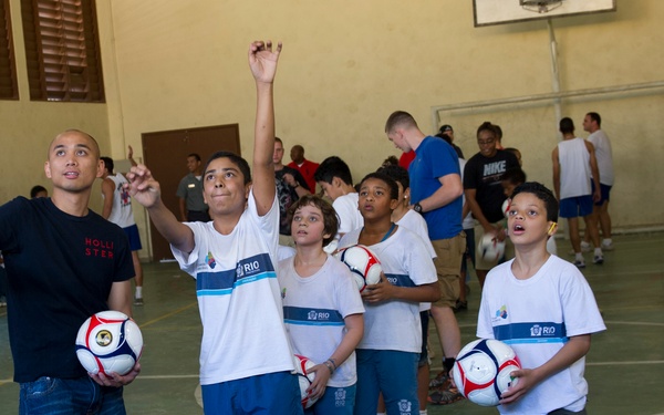 A group of children wearing white 'RIO' branded uniforms are participating in an indoor soccer training session in a gymnasium. An adult instructor in a black Hollister shirt holds a soccer ball while a young boy points upward, apparently tracking a ball or demonstrating a technique. The setting appears to be a community sports program in Rio de Janeiro, Brazil.