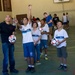 A group of children wearing white 'RIO' branded uniforms are participating in an indoor soccer training session in a gymnasium. An adult instructor in a black Hollister shirt holds a soccer ball while a young boy points upward, apparently tracking a ball or demonstrating a technique. The setting appears to be a community sports program in Rio de Janeiro, Brazil.