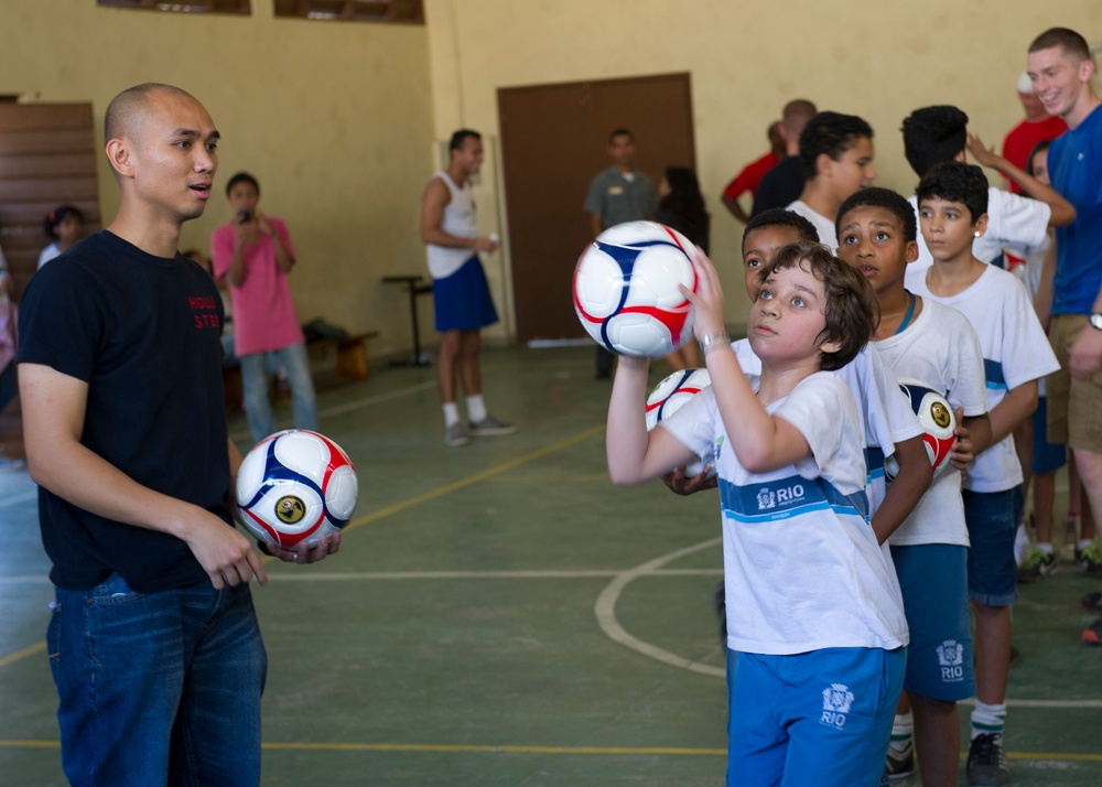 A soccer training session is taking place in an indoor gymnasium, where an adult instructor in a black shirt is working with a group of children wearing white 'RIO' branded uniforms. A child in the foreground is holding a soccer ball overhead, appearing to practice a throw-in technique. The setting appears to be a community sports program in Rio de Janeiro, Brazil.