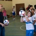 A soccer training session is taking place in an indoor gymnasium, where an adult instructor in a black shirt is working with a group of children wearing white 'RIO' branded uniforms. A child in the foreground is holding a soccer ball overhead, appearing to practice a throw-in technique. The setting appears to be a community sports program in Rio de Janeiro, Brazil.