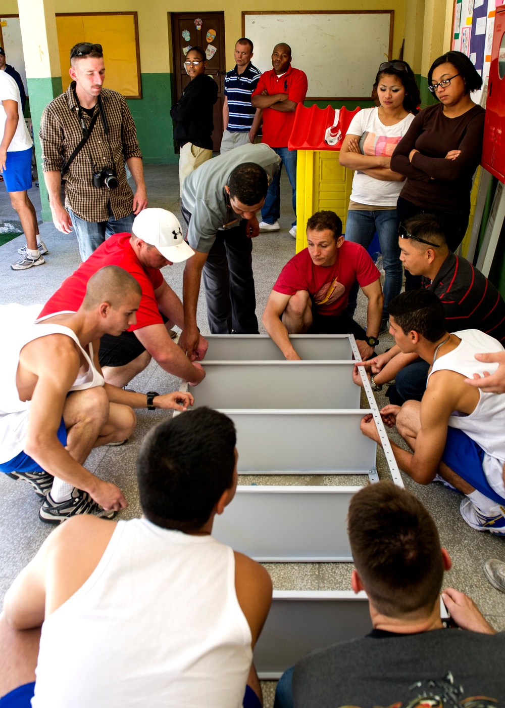 A group of people, many in athletic wear, are assembled inside what appears to be a school or community building, working together to assemble a large metal shelving unit or furniture piece on the floor. Several individuals are crouched down handling the gray metal frame while others stand and observe. The setting appears to be a volunteer or community service activity.