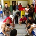 A group of people, many in athletic wear, are assembled inside what appears to be a school or community building, working together to assemble a large metal shelving unit or furniture piece on the floor. Several individuals are crouched down handling the gray metal frame while others stand and observe. The setting appears to be a volunteer or community service activity.