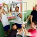 A group of five men are collaboratively assembling a metal shelving unit inside what appears to be a school or community center. The team includes individuals in casual and athletic clothing, working together at various positions around the shelf structure. The setting features colorful wall decorations and murals in the background.