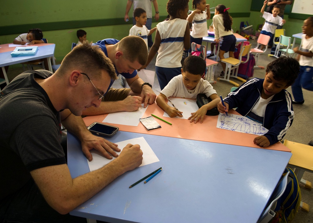A classroom scene showing two adult males sitting at a blue table with two young boys, all engaged in drawing or writing activities on white paper. The room is filled with children in what appears to be school uniforms (white shirts with blue accents), suggesting this is an elementary school setting. The adults appear to be volunteers or visitors participating in an art or educational activity with the students.