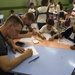 A classroom scene showing two adult males sitting at a blue table with two young boys, all engaged in drawing or writing activities on white paper. The room is filled with children in what appears to be school uniforms (white shirts with blue accents), suggesting this is an elementary school setting. The adults appear to be volunteers or visitors participating in an art or educational activity with the students.