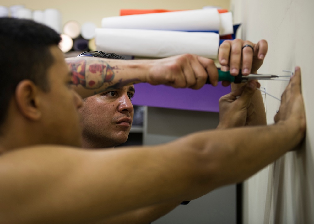 A shirtless man with a colorful tattoo on his forearm is using a screwdriver to work on a wall, appearing to install or adjust a fixture or hook. He is focused on the task, pressing one hand against the wall for support while driving a screw. The background shows shelves with rolled materials and a purple accent wall.