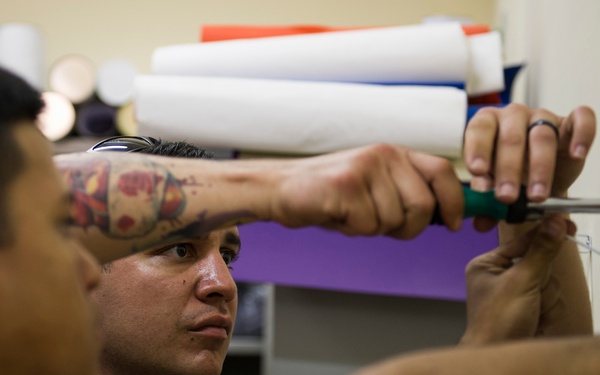 A shirtless man with a colorful tattoo on his forearm is using a screwdriver to work on a wall, appearing to install or adjust a fixture or hook. He is focused on the task, pressing one hand against the wall for support while driving a screw. The background shows shelves with rolled materials and a purple accent wall.