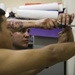 A shirtless man with a colorful tattoo on his forearm is using a screwdriver to work on a wall, appearing to install or adjust a fixture or hook. He is focused on the task, pressing one hand against the wall for support while driving a screw. The background shows shelves with rolled materials and a purple accent wall.
