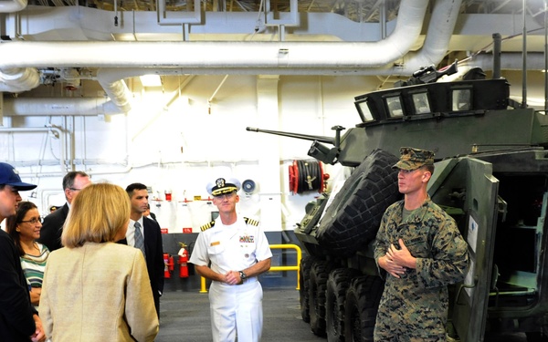 A U.S. Marine in camouflage utilities is briefing a group of civilian visitors and a Navy officer in dress whites inside what appears to be the well deck or vehicle bay of a naval vessel. A large wheeled armored vehicle, consistent with an LAV-25 (Light Armored Vehicle), is secured with chains in the background. The setting suggests an official tour or congressional/VIP visit to a U.S. Navy amphibious ship.
