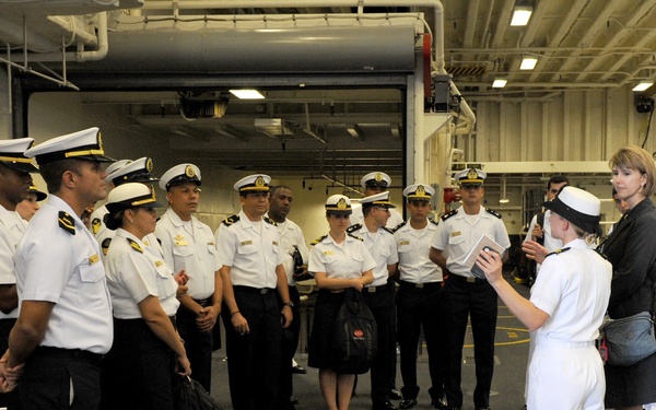 A group of naval officers in white dress uniforms with gold insignia are gathered in what appears to be the hangar bay of a naval vessel. A female officer in white uniform and a civilian woman appear to be briefing or conducting a tour for the assembled group. The setting suggests an official visit or orientation aboard a military ship.
