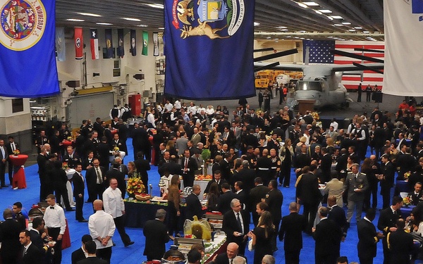 A large formal reception or military gala is being held inside what appears to be a naval vessel's hangar bay, identifiable by the industrial ceiling structure and a visible helicopter in the background. Hundreds of attendees dressed in formal civilian attire and military dress uniforms mingle around food and beverage stations on a blue carpeted floor. Multiple state and organizational flags are prominently displayed overhead, suggesting an official government or military ceremonial event.