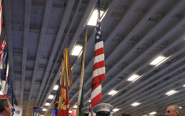 A U.S. Marine Corps Color Guard is marching in a formal ceremony inside a large indoor venue with a blue carpet aisle. The lead Marine carries the U.S. national flag while others carry additional military and organizational flags. Attendees in military dress uniforms and civilian attire stand at attention on both sides of the aisle.