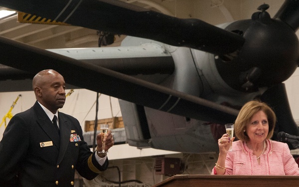 A naval officer in dress uniform and a woman in a pink blazer are raising glasses in a toast during a formal ceremony. The event appears to be taking place inside a ship's hangar bay, with a vintage propeller aircraft visible in the background. The woman is standing at a wooden podium with a microphone, suggesting she may be delivering remarks.