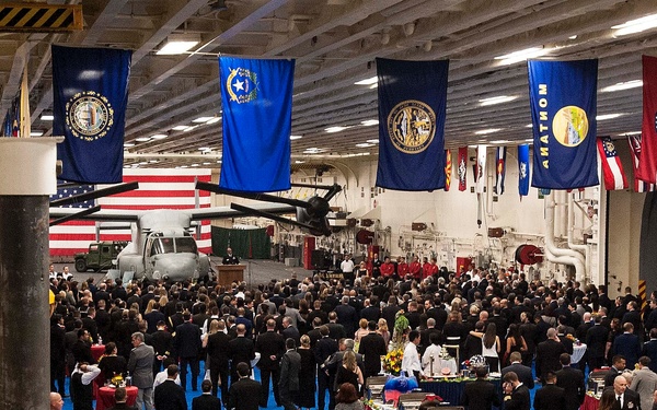 A formal military reception or gala event is being held inside the hangar bay of a naval aircraft carrier. Hundreds of attendees dressed in formal attire and military dress uniforms mingle around buffet tables set with red tablecloths. The space is decorated with U.S. state flags hanging from the overhead structure, and a military rotary-wing aircraft (appears to be an MV-22 Osprey or similar) is visible in the background.