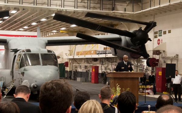 A formal military ceremony is taking place inside a large hangar bay, likely aboard a naval vessel or military installation. A uniformed officer is delivering a speech at a wooden podium before a crowd of attendees in civilian and military attire. A V-22 Osprey tiltrotor aircraft and a military vehicle are prominently displayed in the background, with a large American flag hanging on the wall.
