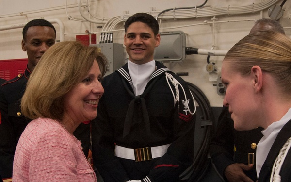A civilian woman in a pink tweed suit is shaking hands with a female military officer in dress uniform with white aiguillette. They are surrounded by several service members in formal dress uniforms, including U.S. Marines in dress blues with white gloves and gold belts, and a U.S. Navy sailor in dress uniform. The setting appears to be aboard a naval vessel or military facility.